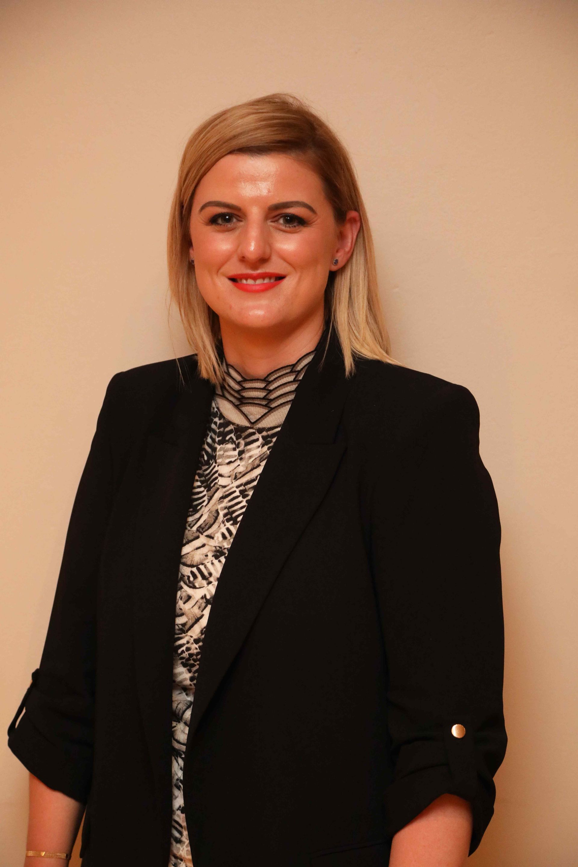 Woman in a black blazer standing against a beige wall, smiling at the camera.