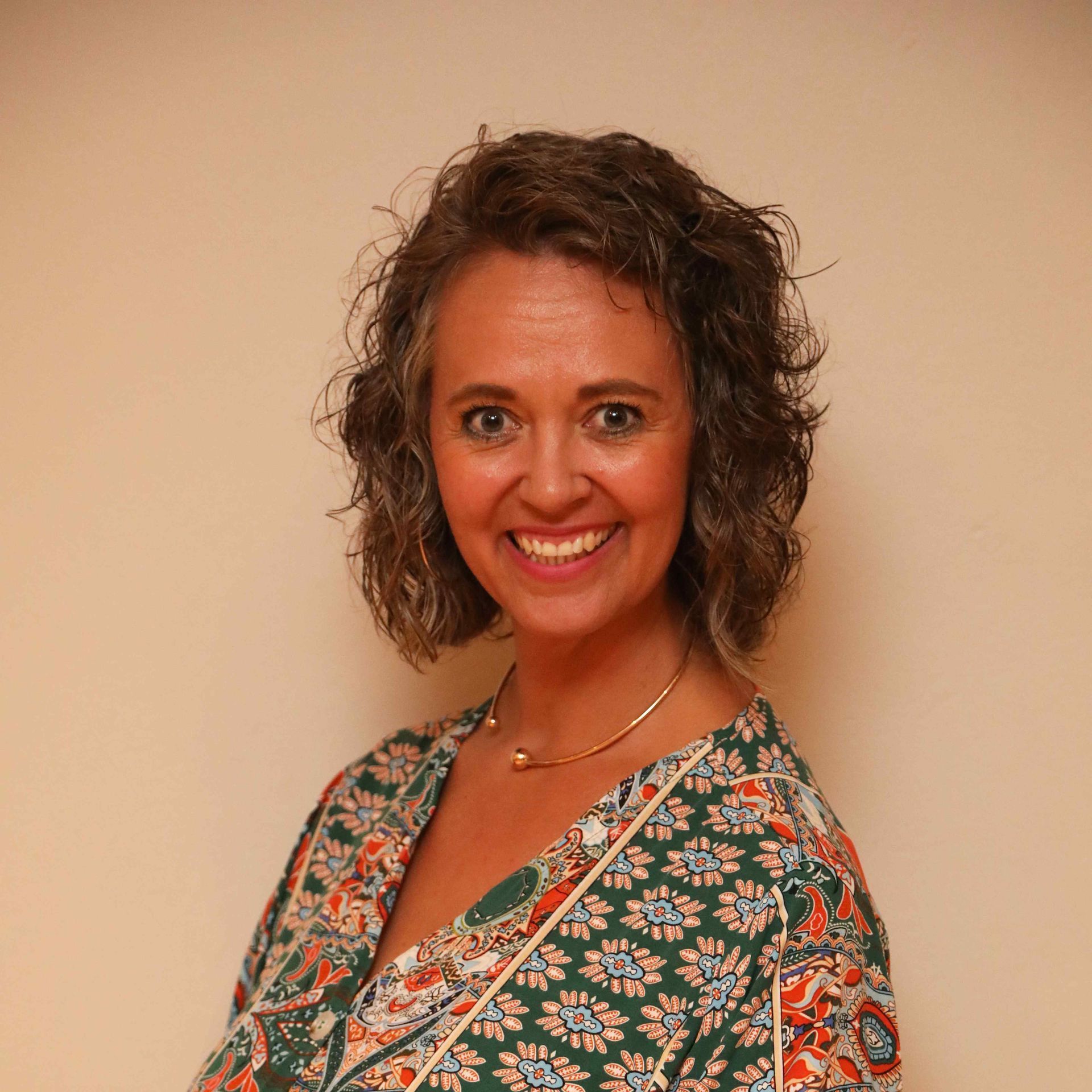 Smiling woman with curly hair in a green floral top against a beige wall
