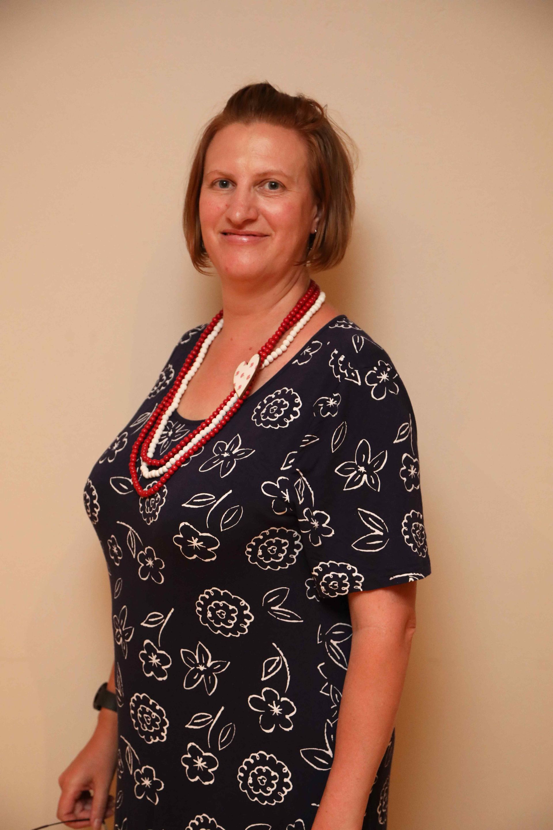 Woman standing against a beige wall, wearing a navy floral dress and layered red-white necklace.