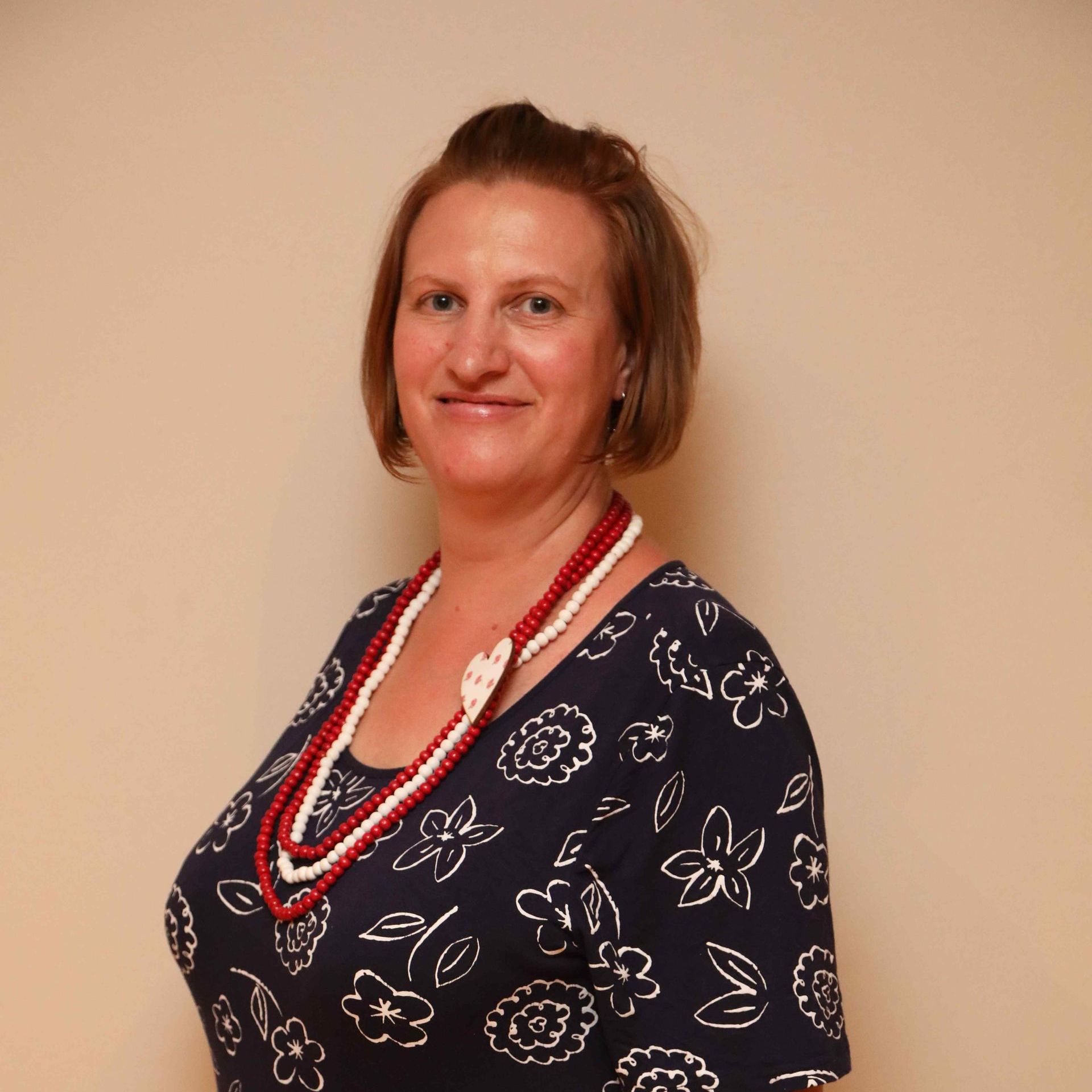Portrait of a smiling person in a navy floral top and red necklace against a beige background
