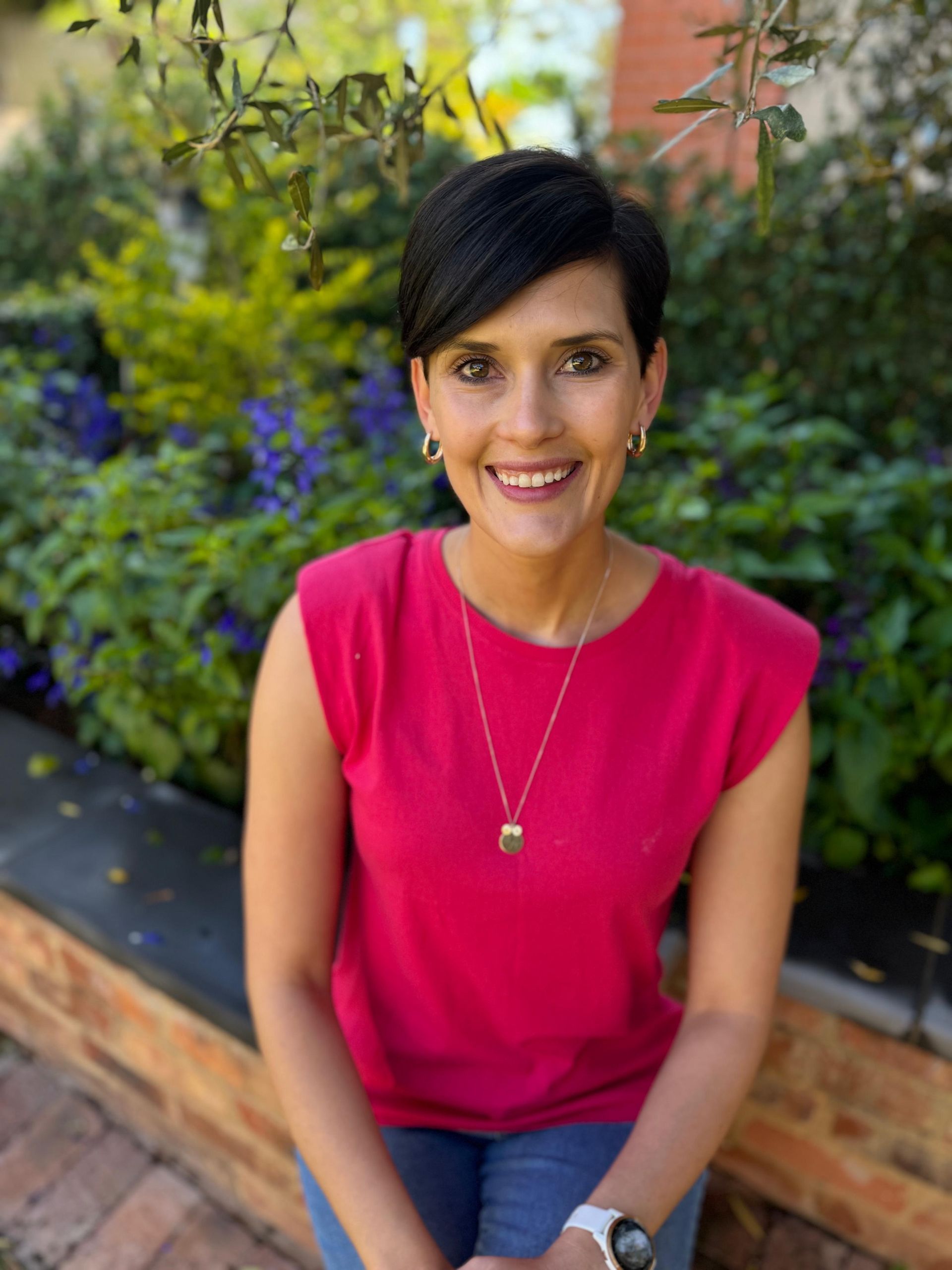 Woman in a bright pink top smiling outdoors beside flowers and greenery