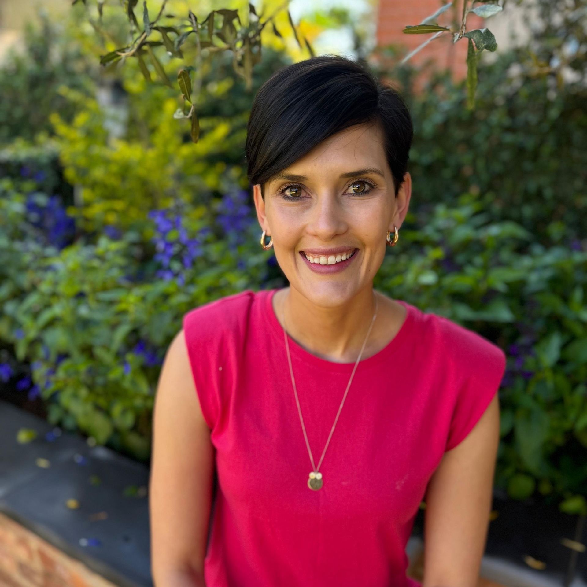 Smiling person in a bright pink top standing outdoors in a garden setting.