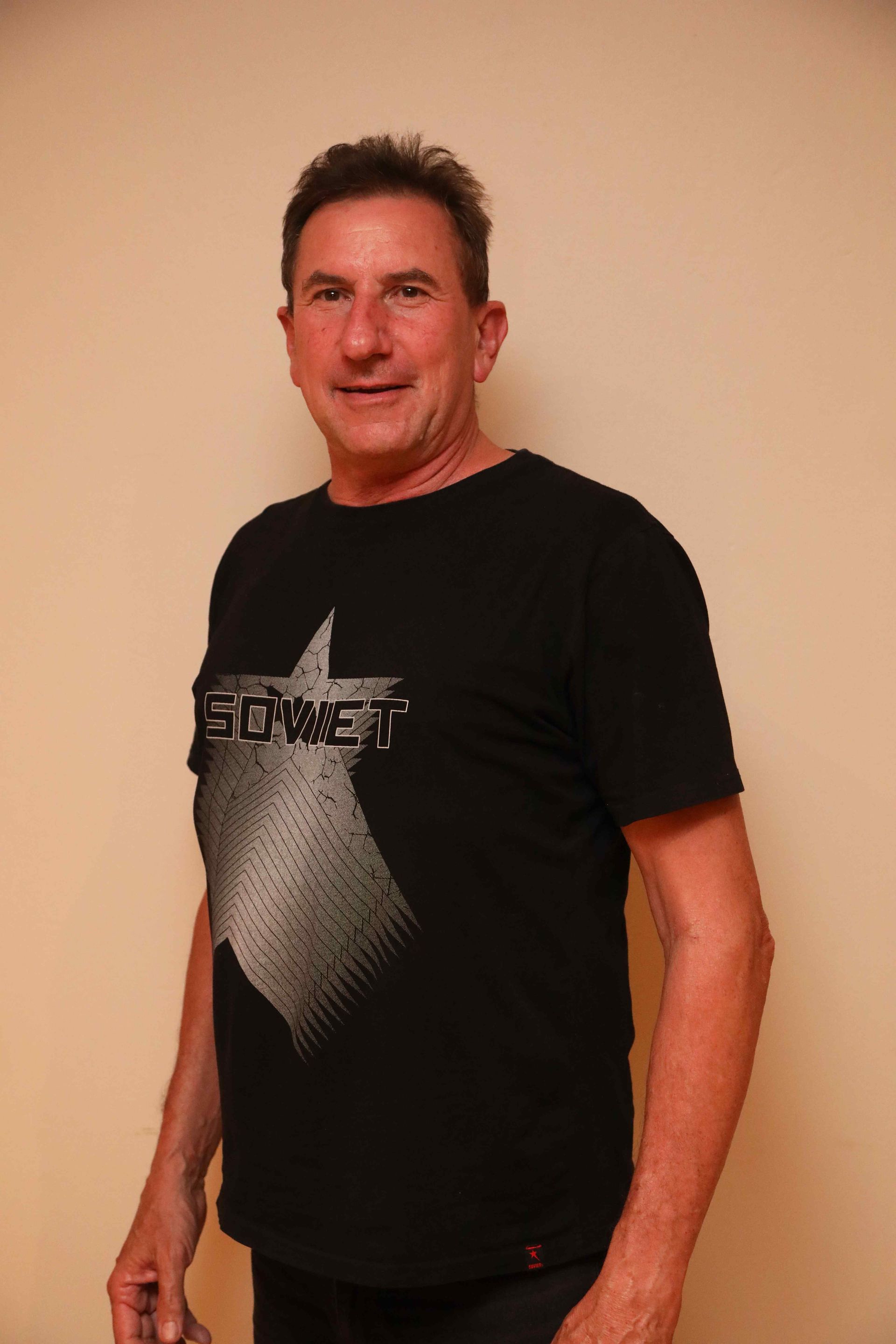 Man standing against a beige wall, wearing a black graphic T-shirt and jeans, smiling slightly.