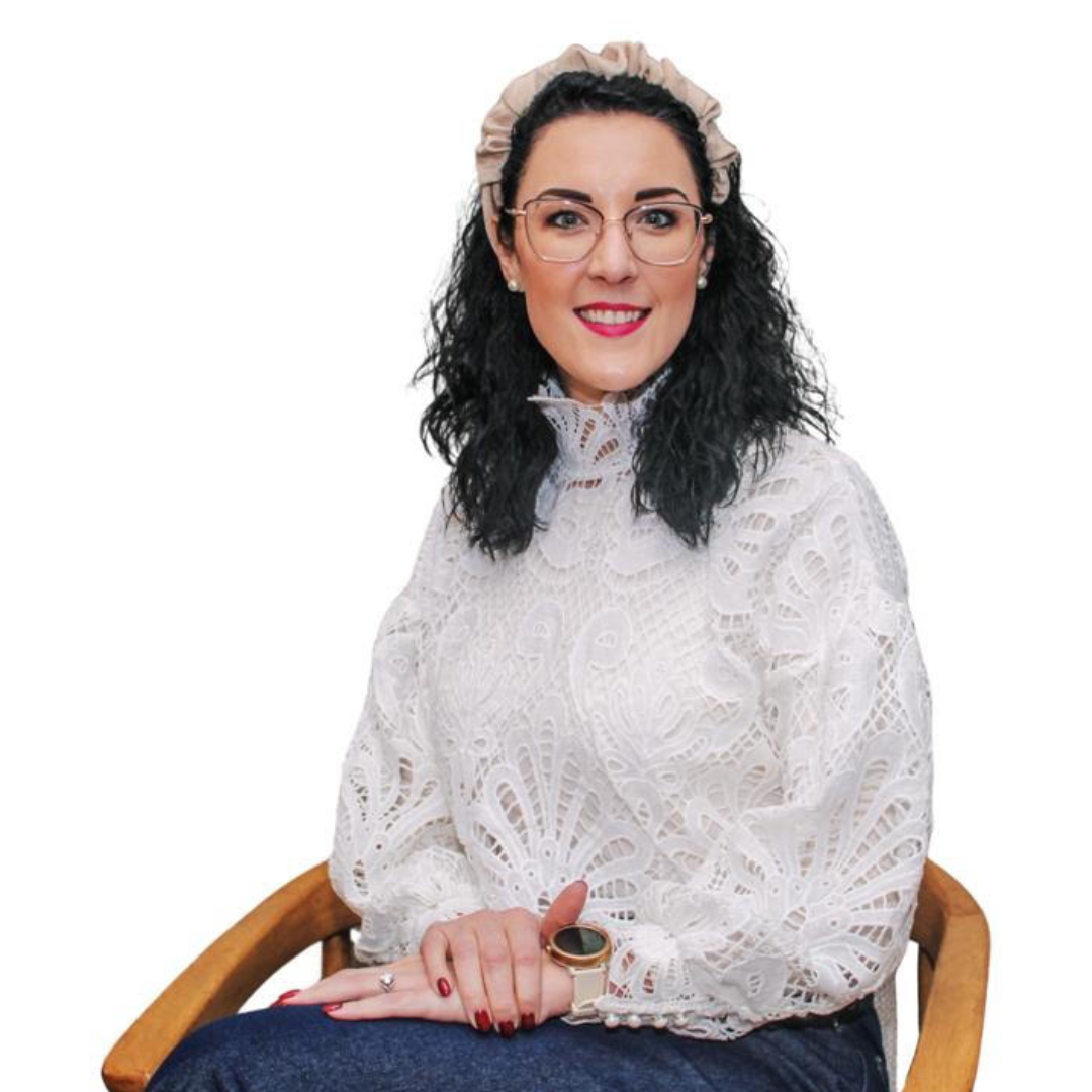 Woman seated in a wooden chair, wearing a white lace blouse and glasses, smiling at the camera.
