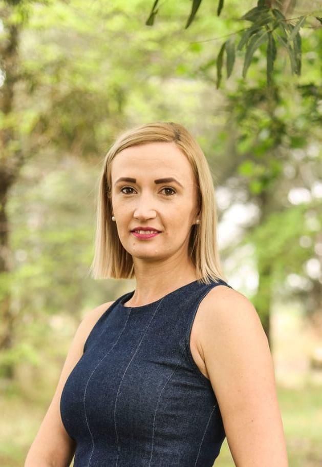 Portrait of a blonde woman in a navy sleeveless dress standing outdoors with green foliage behind her