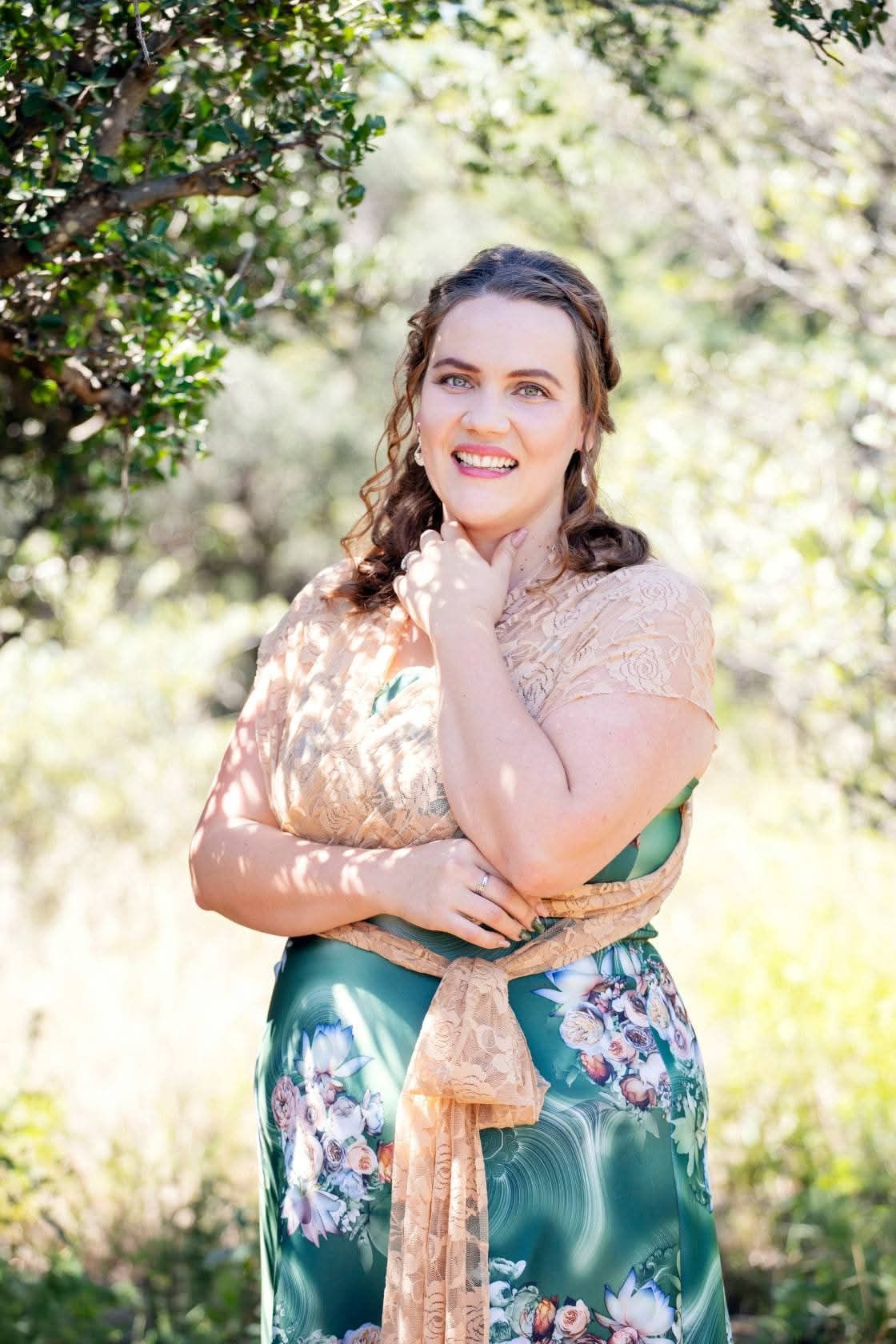 Smiling woman standing outdoors among greenery, wearing a floral dress and shawl.