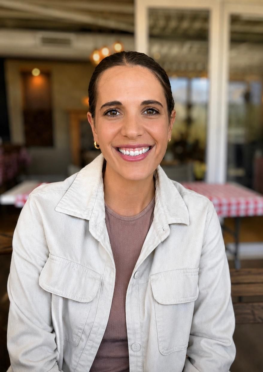 Smiling person in a white jacket seated outdoors at a café with blurred tables in the background