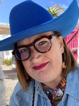 Person in a bright blue wide-brimmed hat and glasses, smiling outdoors in colorful jewelry.