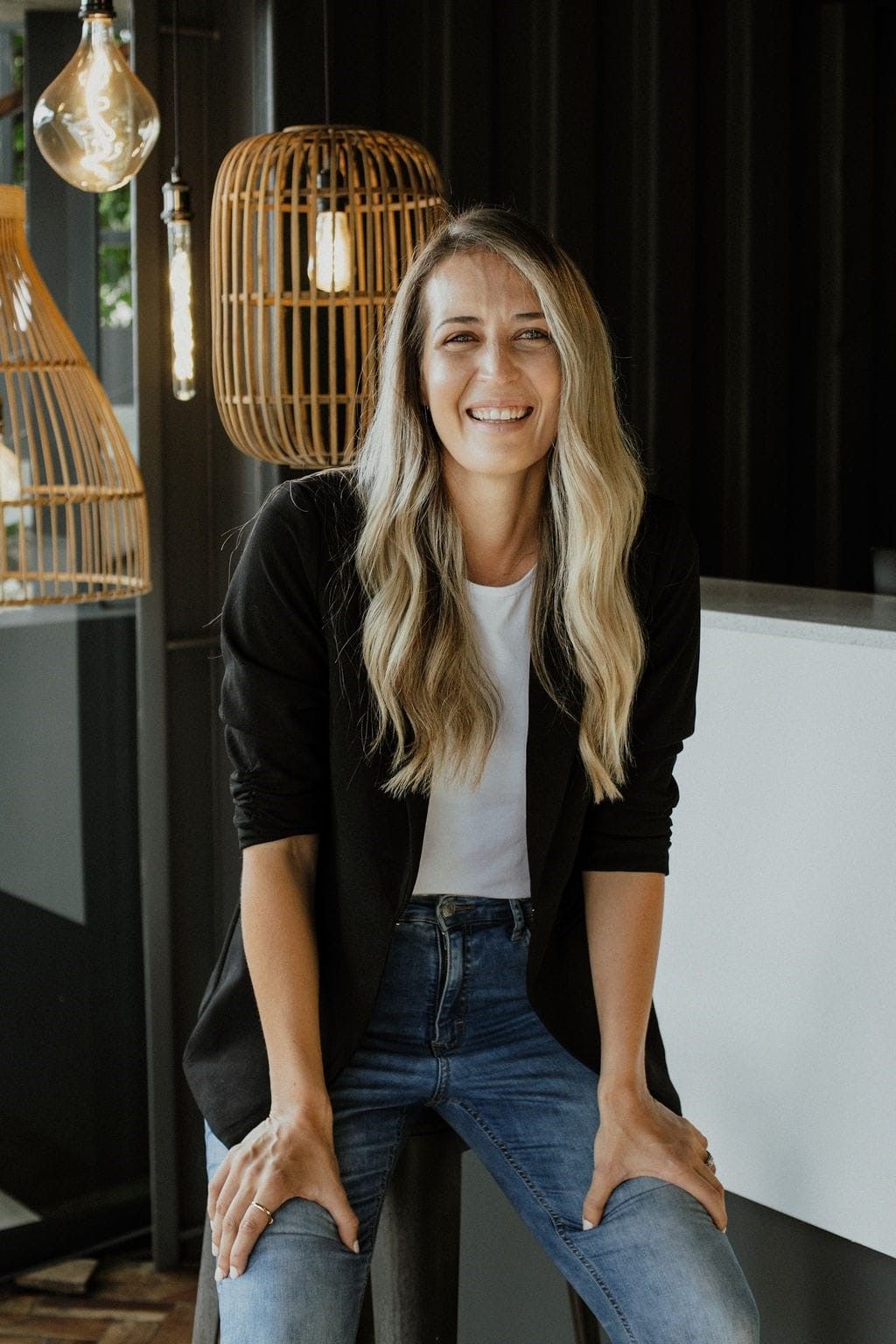 Smiling woman seated on a stool in a modern interior with black walls and woven pendant lights.