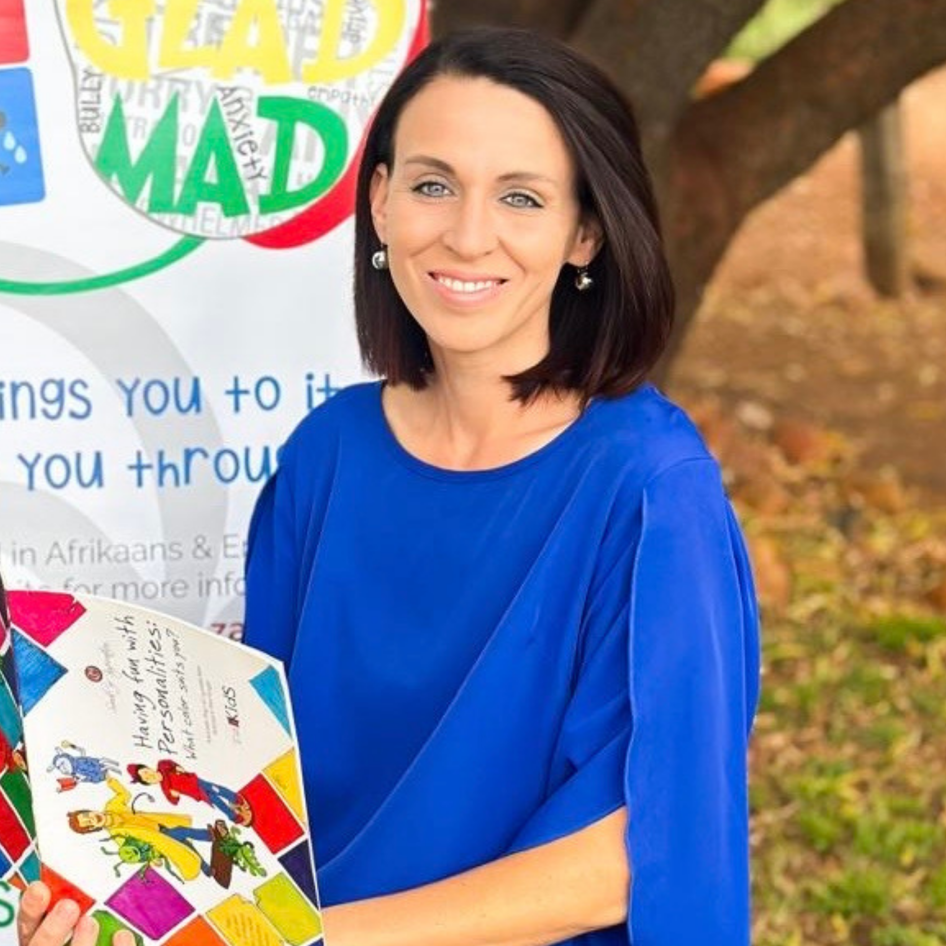 Woman in a blue dress smiling beside a colorful sign outdoors, holding a patterned folder