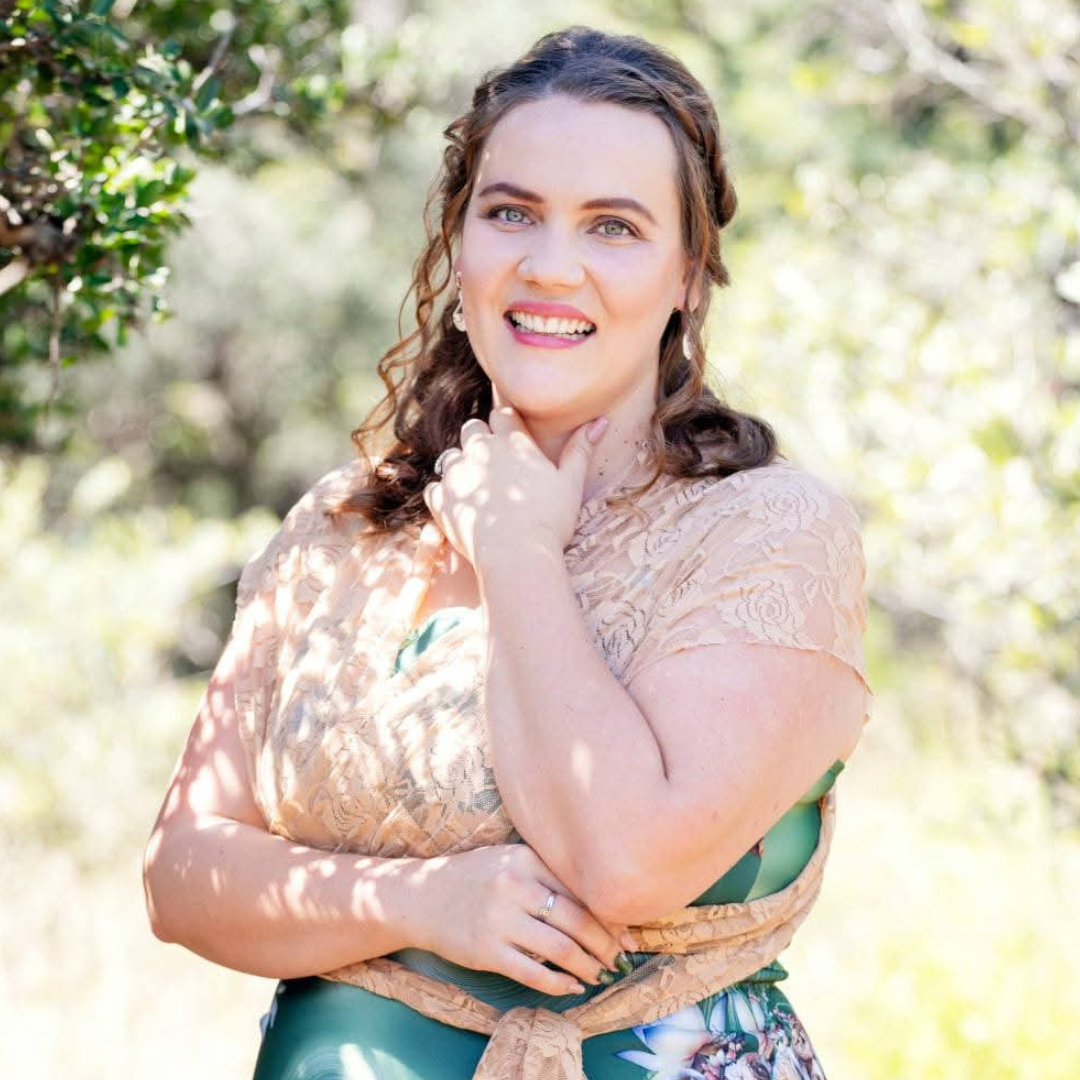 Smiling woman in a lace dress standing outdoors in bright sunlight, with greenery in the background