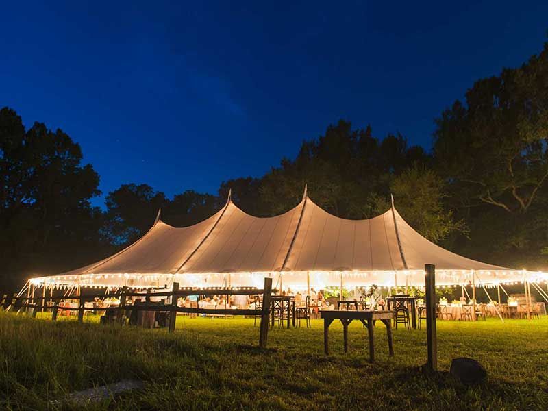 Illuminated outdoor tent at dusk with people gathered inside, surrounded by trees and grass