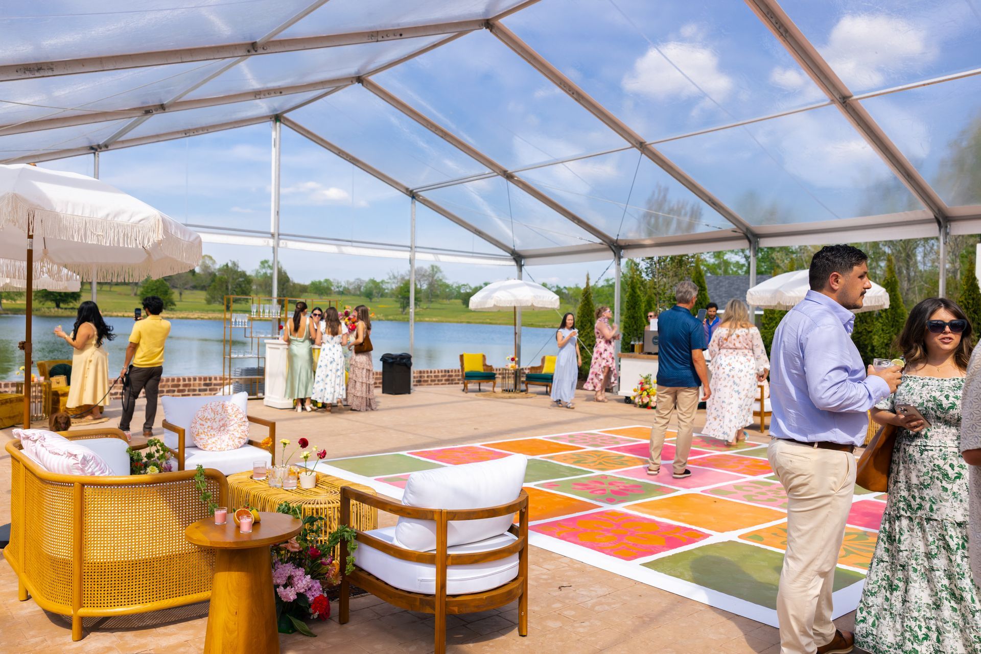 People socializing under a clear tent by a lake, with colorful rugs and tables set for an outdoor event