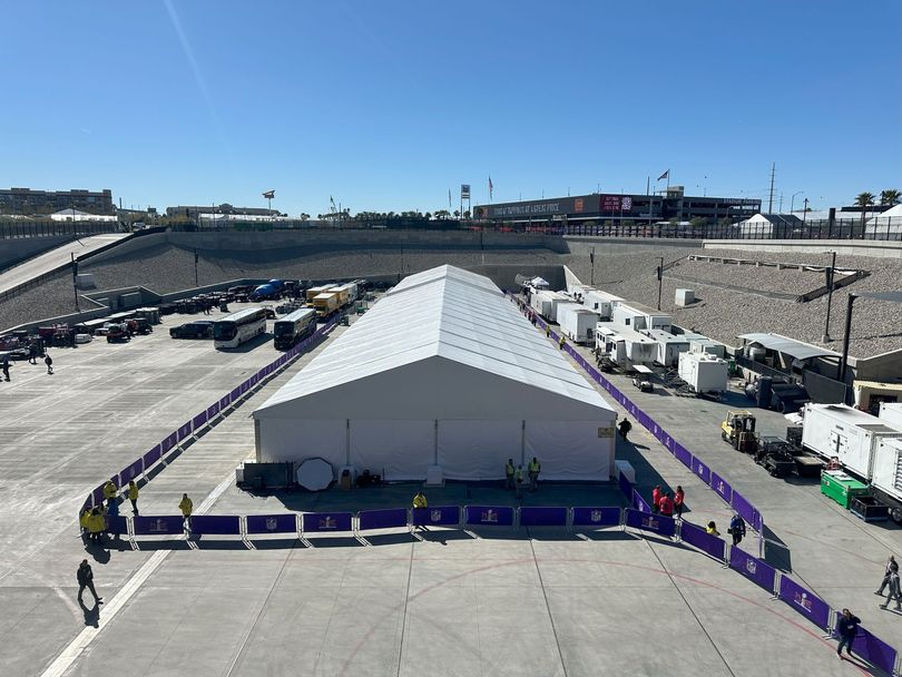 Large white event tent in a fenced paved area, with vehicles and workers around under a clear sky.