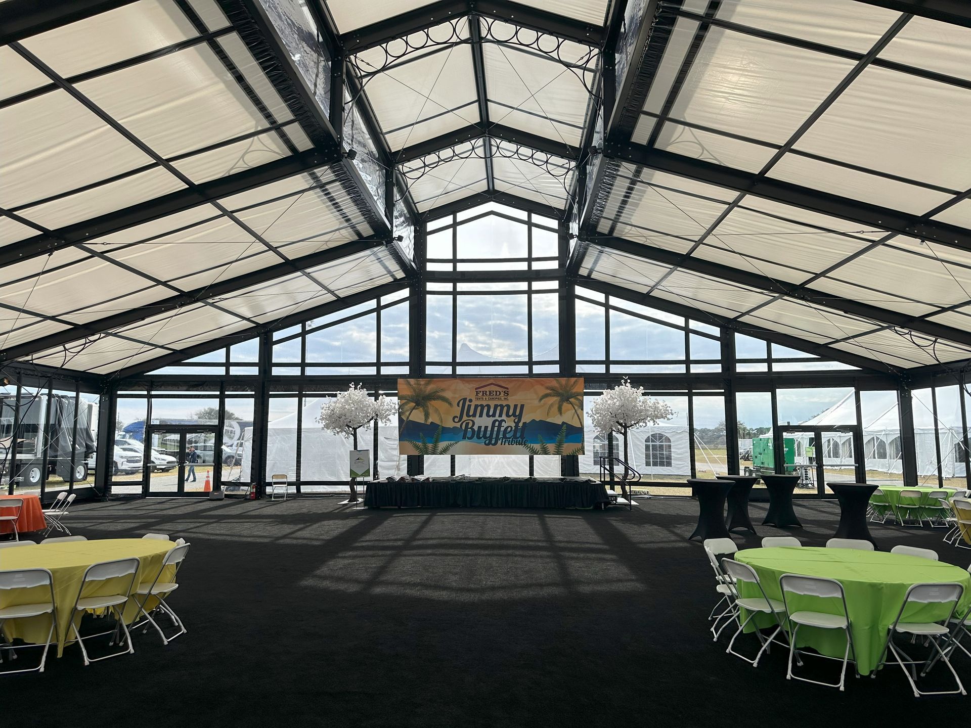 Open-air event pavilion with black floor, glass walls, and round tables dressed in green and yellow linens
