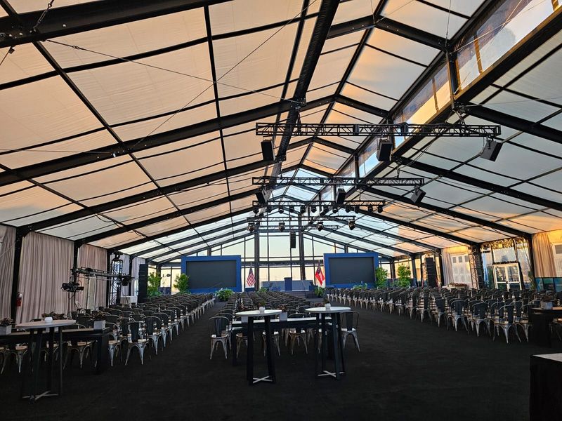 Empty event tent with rows of chairs and tables under a peaked canopy, lit by daylight.