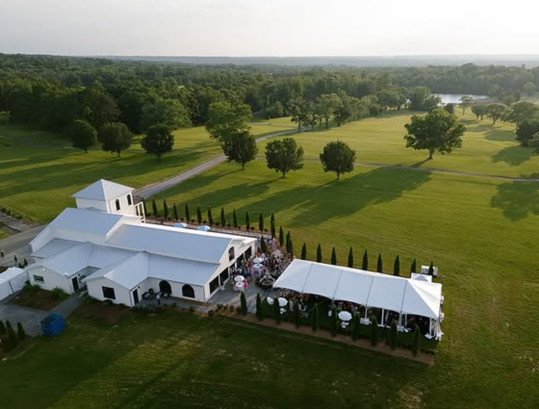 Aerial view of a white event tent beside a pavilion on a green golf course at sunset.