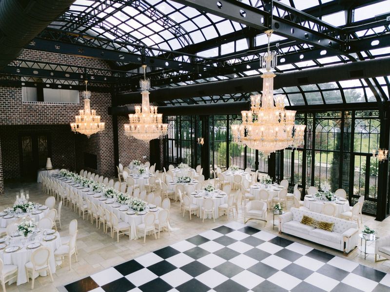 Elegant glass-roofed event hall with white tables, chandeliers, and a black-and-white checkered floor