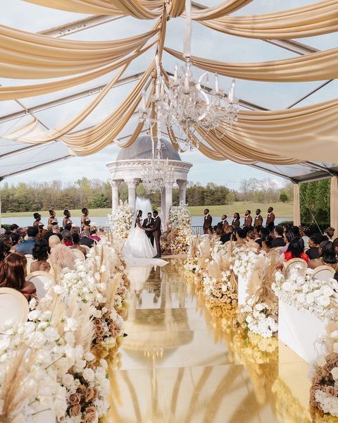 Elegant wedding ceremony under gold drapes with chandeliers, white floral aisle, and mirrored floor