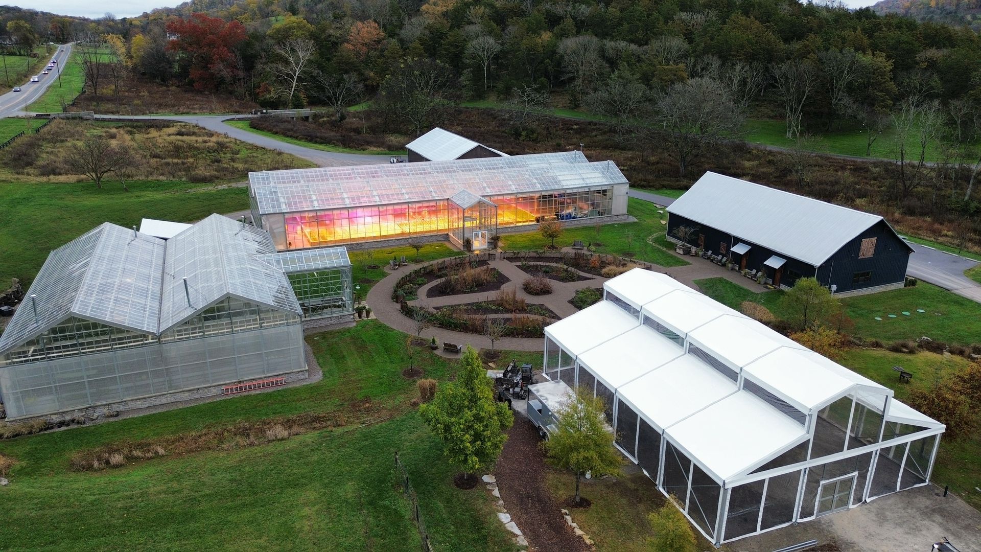 Aerial view of a greenhouse complex with a lit central greenhouse, paths, and surrounding grassy fields.