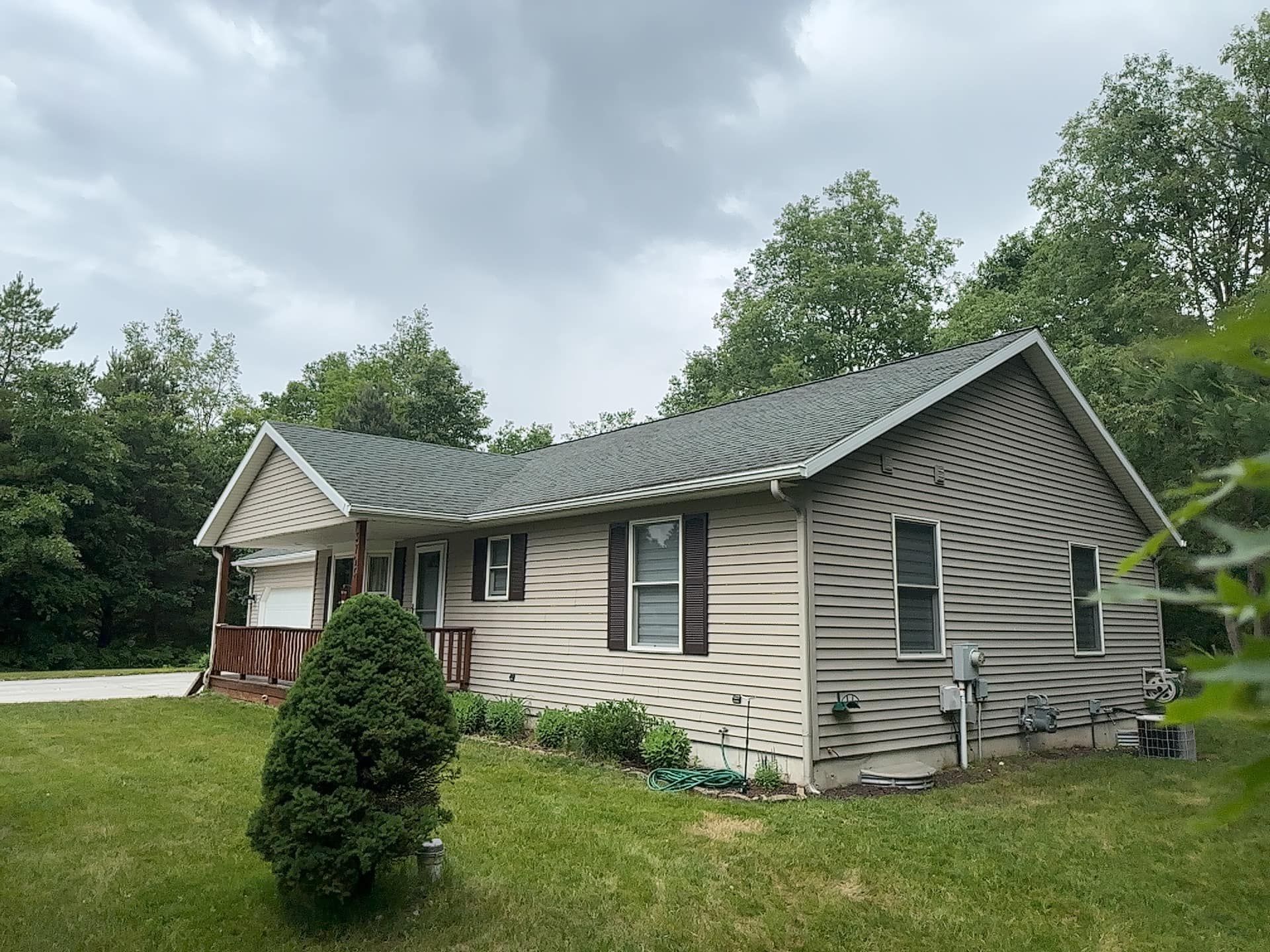 Small beige house with dark roof and siding, surrounded by grass and trees under a cloudy sky