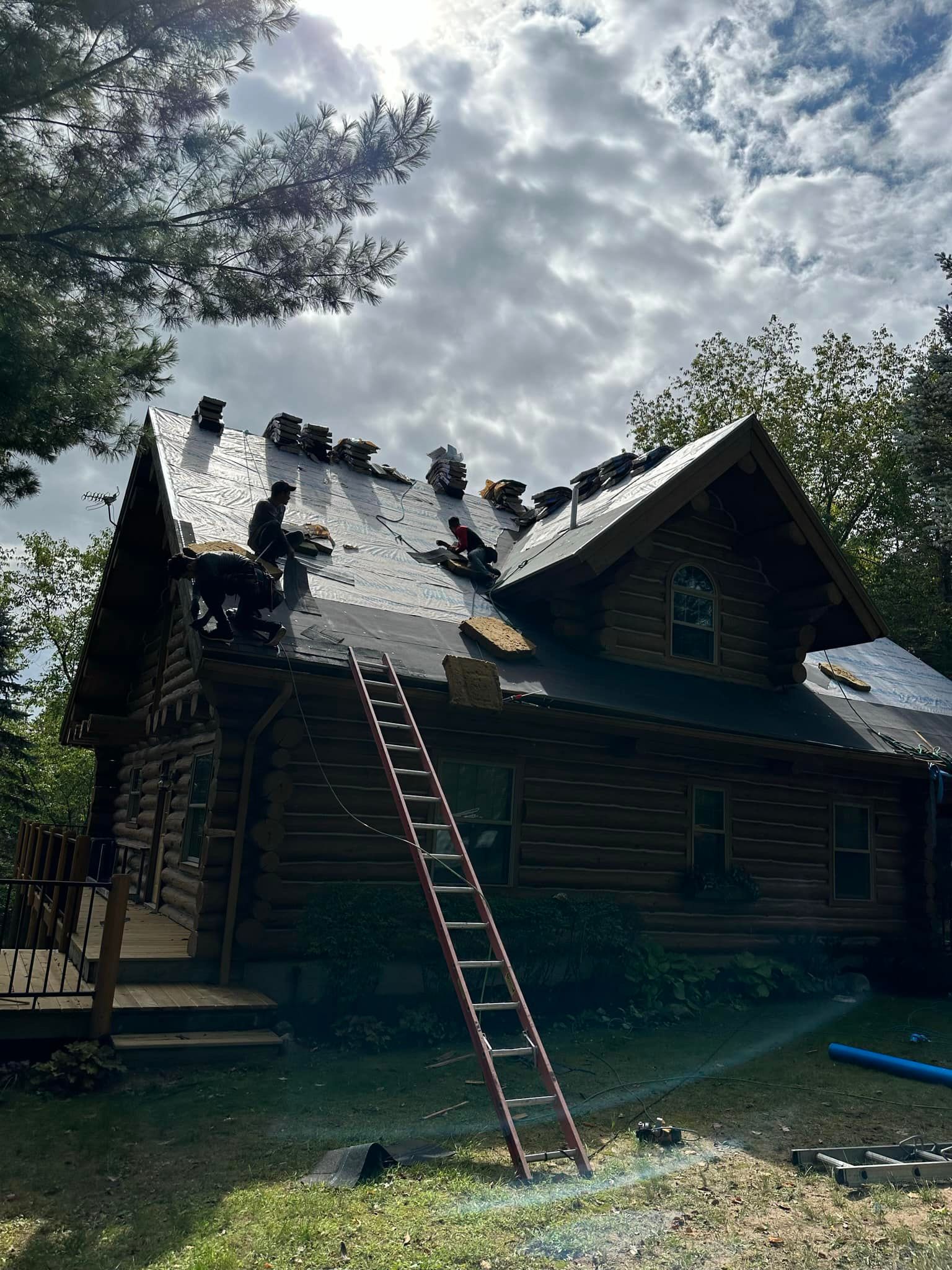 Ladder leaning against a dark wooden cabin roof under a cloudy sky, with workers on the roof.