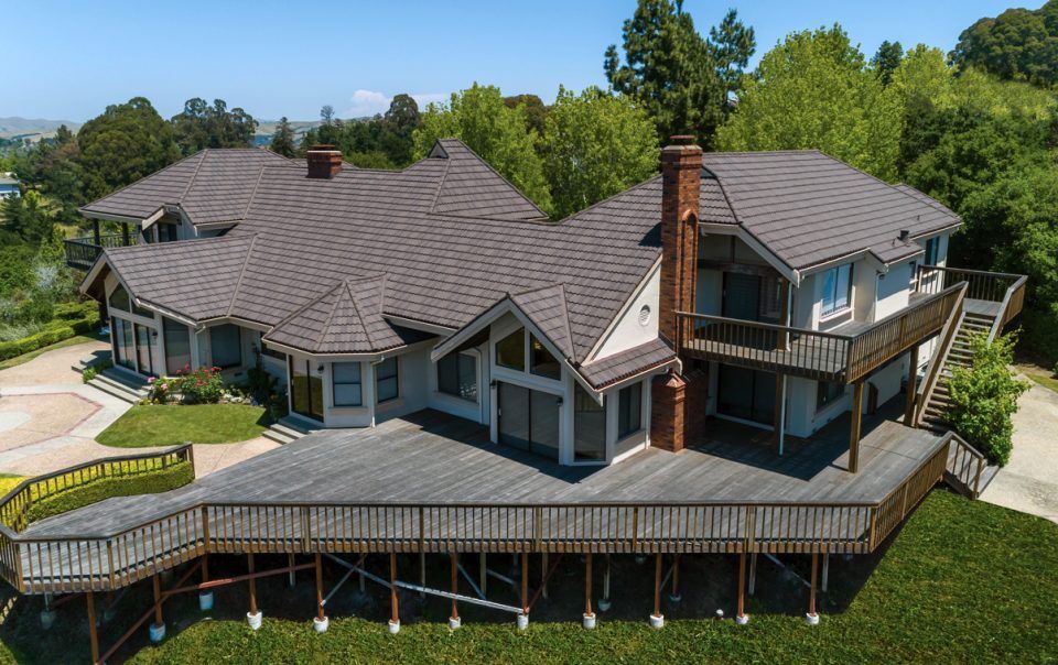 Large house with a gray roof, wraparound deck, and chimney on a grassy hillside surrounded by trees