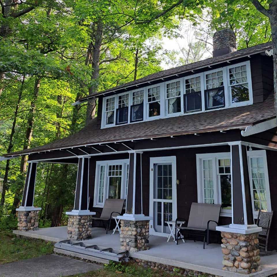Rustic cabin porch with stone columns, white-trimmed windows, and chairs in a wooded setting