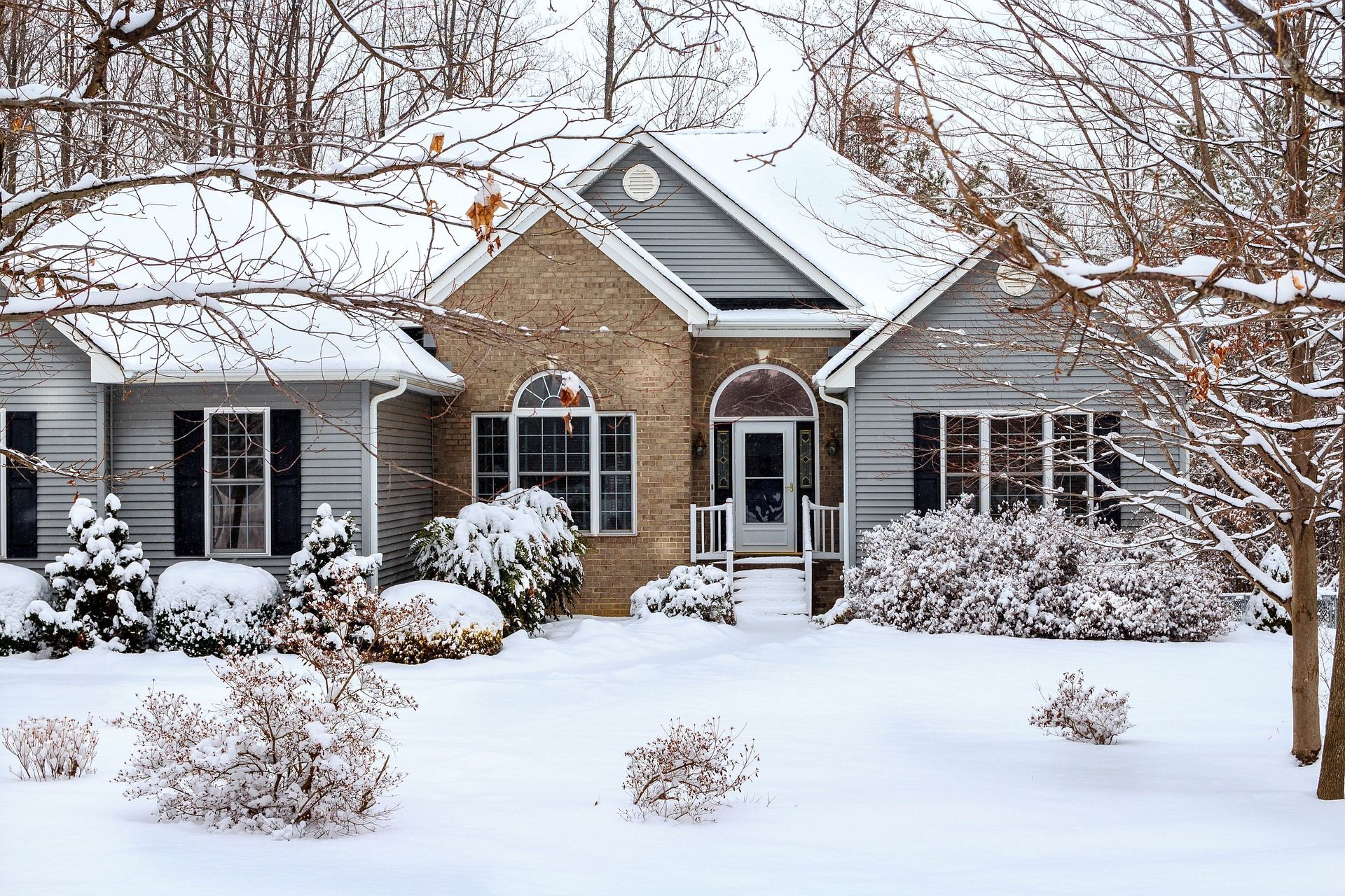 Snow-covered suburban house with brick and gray siding, framed by bare trees and winter shrubs.