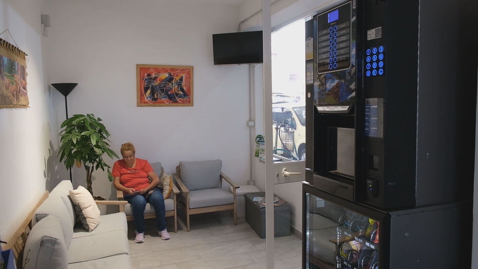 Donna in sala d'attesa vicino a una macchinetta del caffè, seduta su un divano e che guarda il telefono.