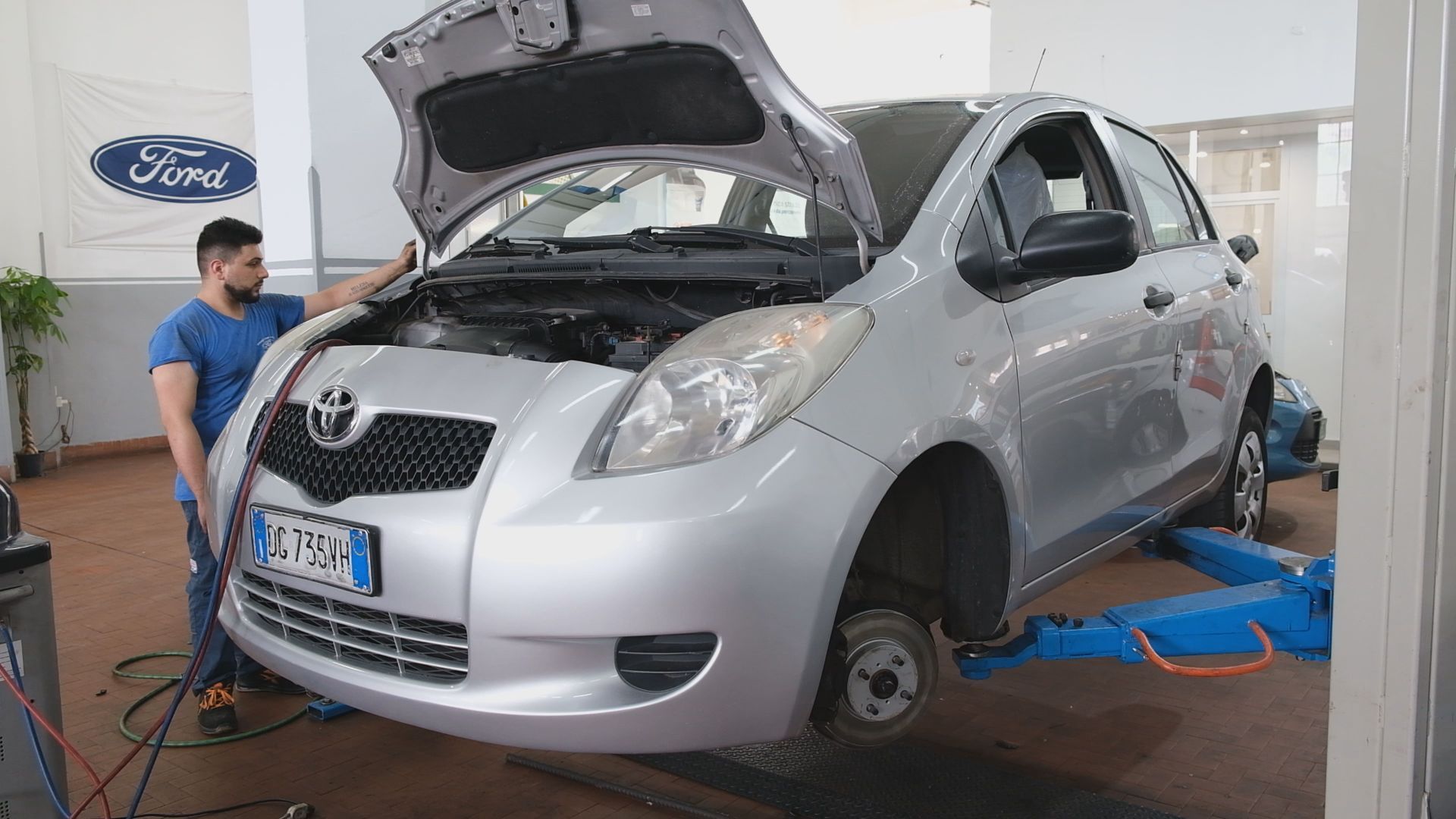 Meccanico con camicia blu al lavoro su una Toyota Yaris argentata, sollevata in un'officina auto a Palermo