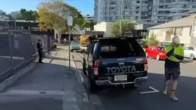 Dark Toyota pickup truck parked; worker in high-vis vest; person standing near a fence. Urban setting, daytime.