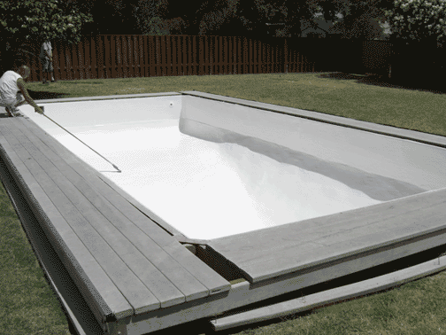 A rectangular above-ground pool with gray decking is being prepared in a grassy backyard, with a person working on the edge.