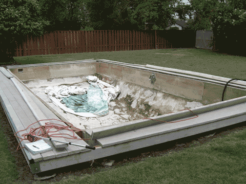 Empty concrete swimming pool in backyard, with wooden deck around it.