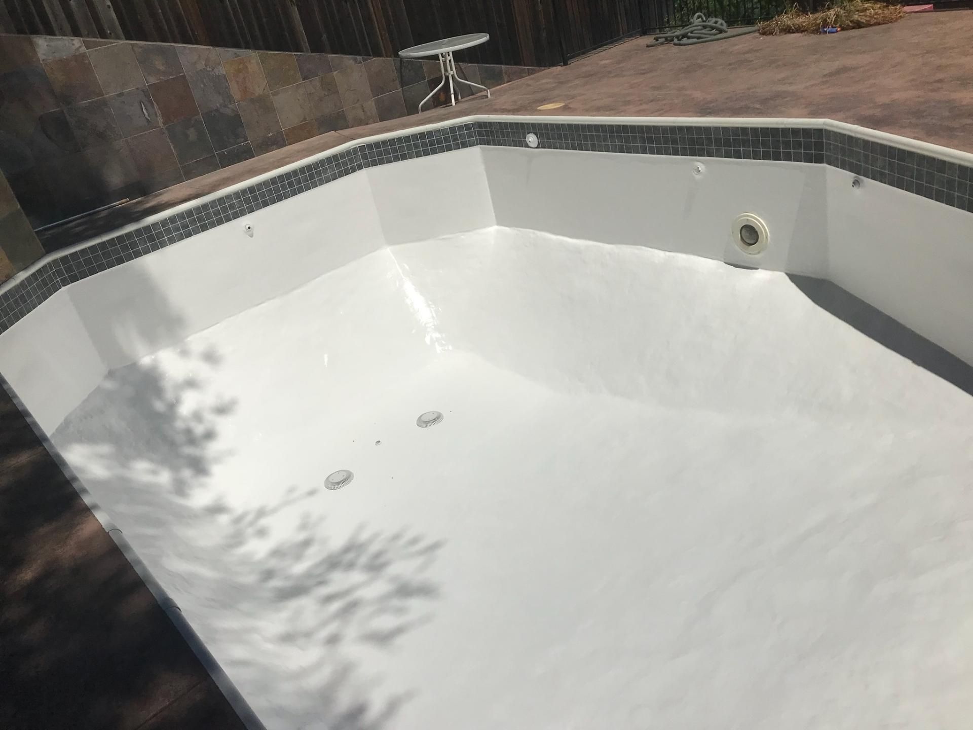 Empty, white-painted swimming pool. Dark tile border, brown patio, and wooden fence surround the pool. Empty, white-painted swimming pool. Dark tile border, brown patio, and wooden fence surround the pool.