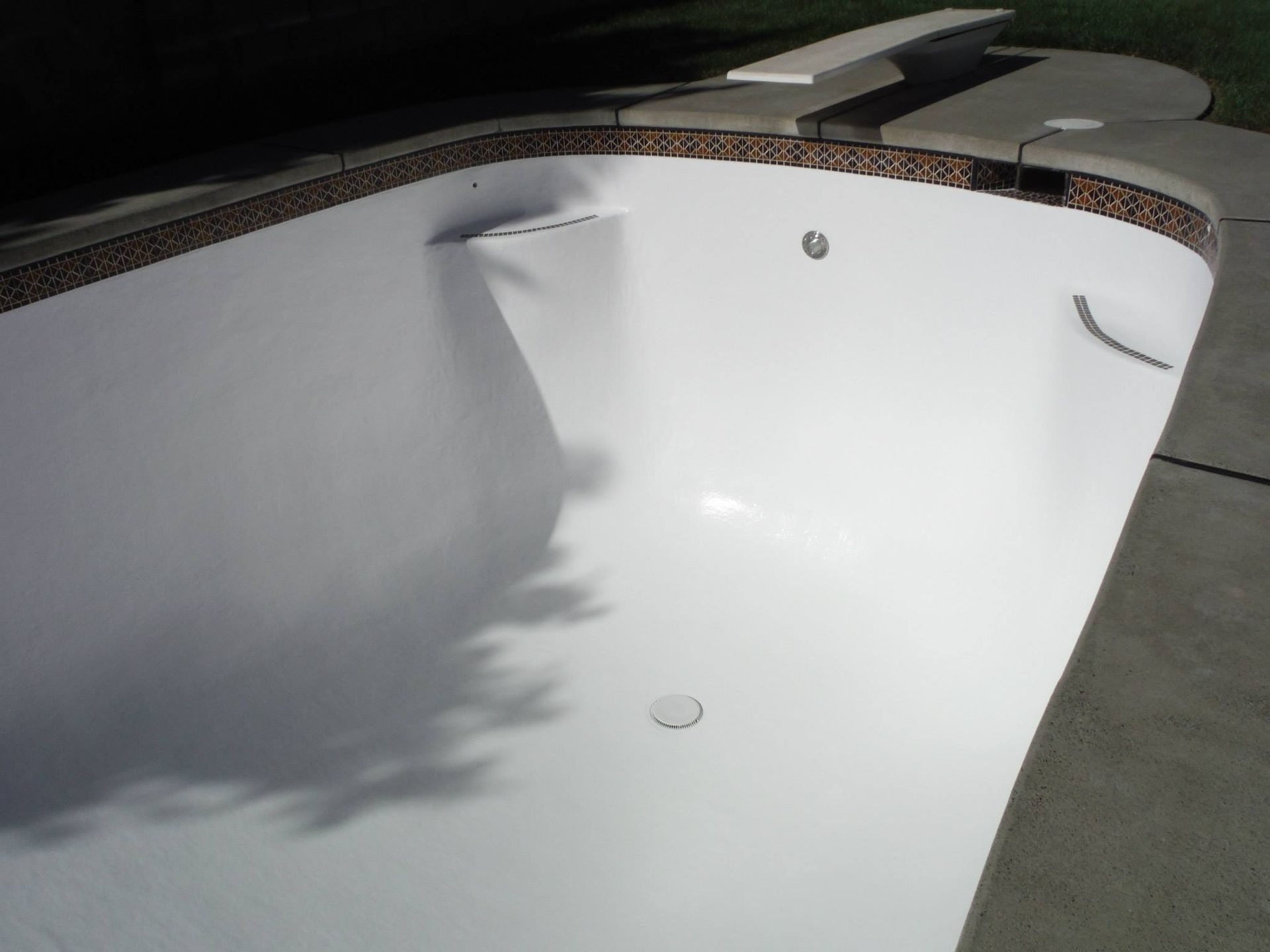Empty white swimming pool with brown tile border and diving board. Concrete deck surrounds. Empty white swimming pool with brown tile border and diving board. Concrete deck surrounds.