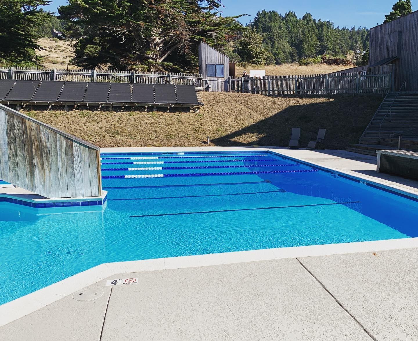 Outdoor swimming pool with blue water. Concrete deck and wooden fence surround. Sunny day. Outdoor swimming pool with blue water. Concrete deck and wooden fence surround. Sunny day.