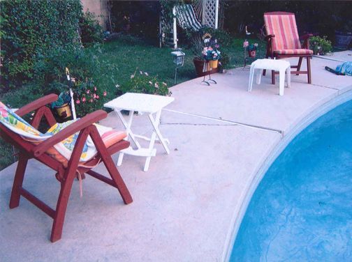 Poolside scene with chairs and small tables on a concrete deck, overlooking a swimming pool and grass.