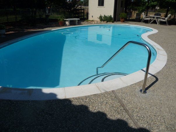 Pool with curved edges, blue water, stainless steel ladder, white coping, and concrete patio. Pool with curved edges, blue water, stainless steel ladder, white coping, and concrete patio.