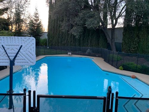 Swimming pool with basketball hoop and surrounding fence, trees in background, during daytime. Swimming pool with basketball hoop and surrounding fence, trees in background, during daytime.