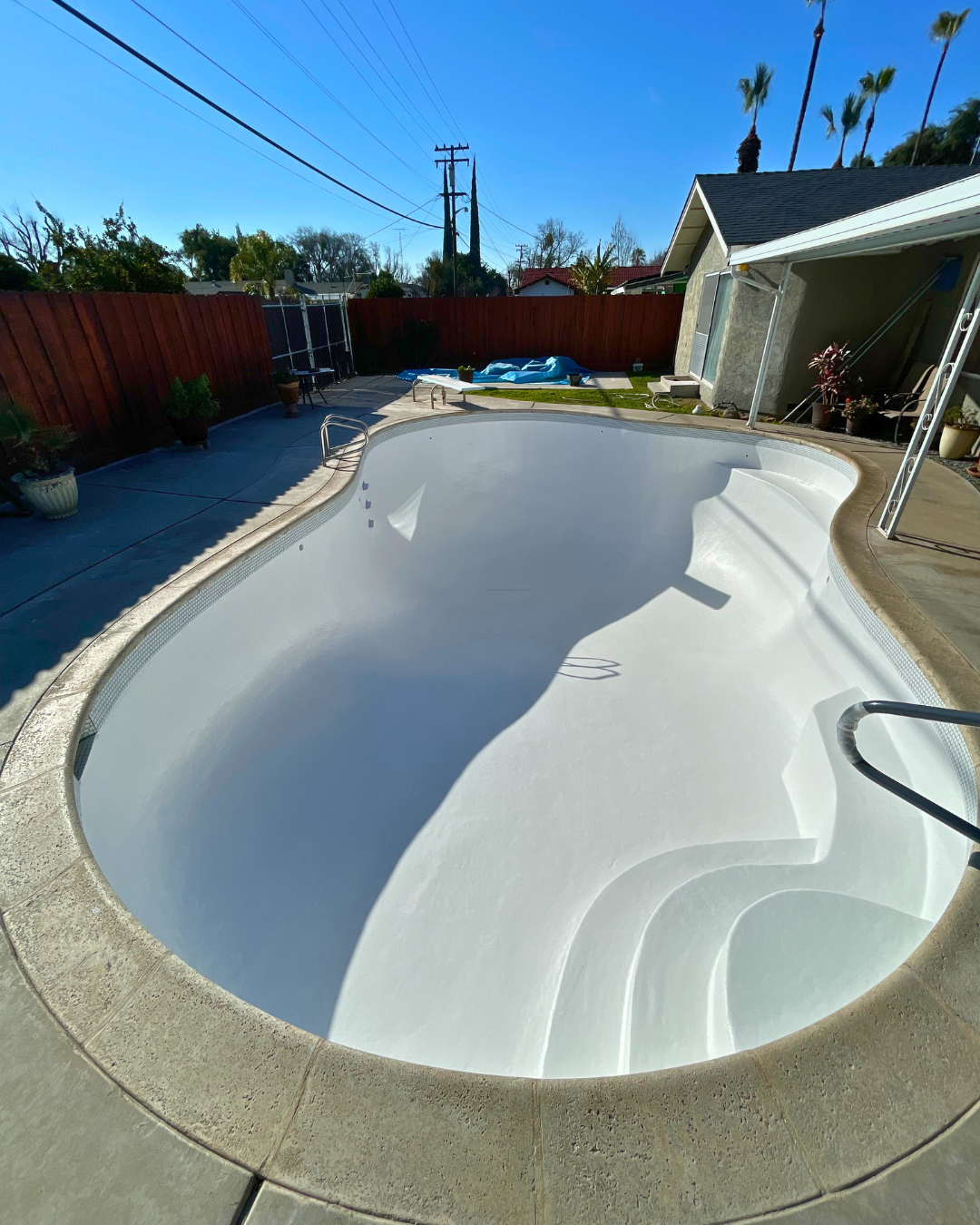 Pool with curved edges, blue water, stainless steel ladder, white coping, and concrete patio. Pool with curved edges, blue water, stainless steel ladder, white coping, and concrete patio.