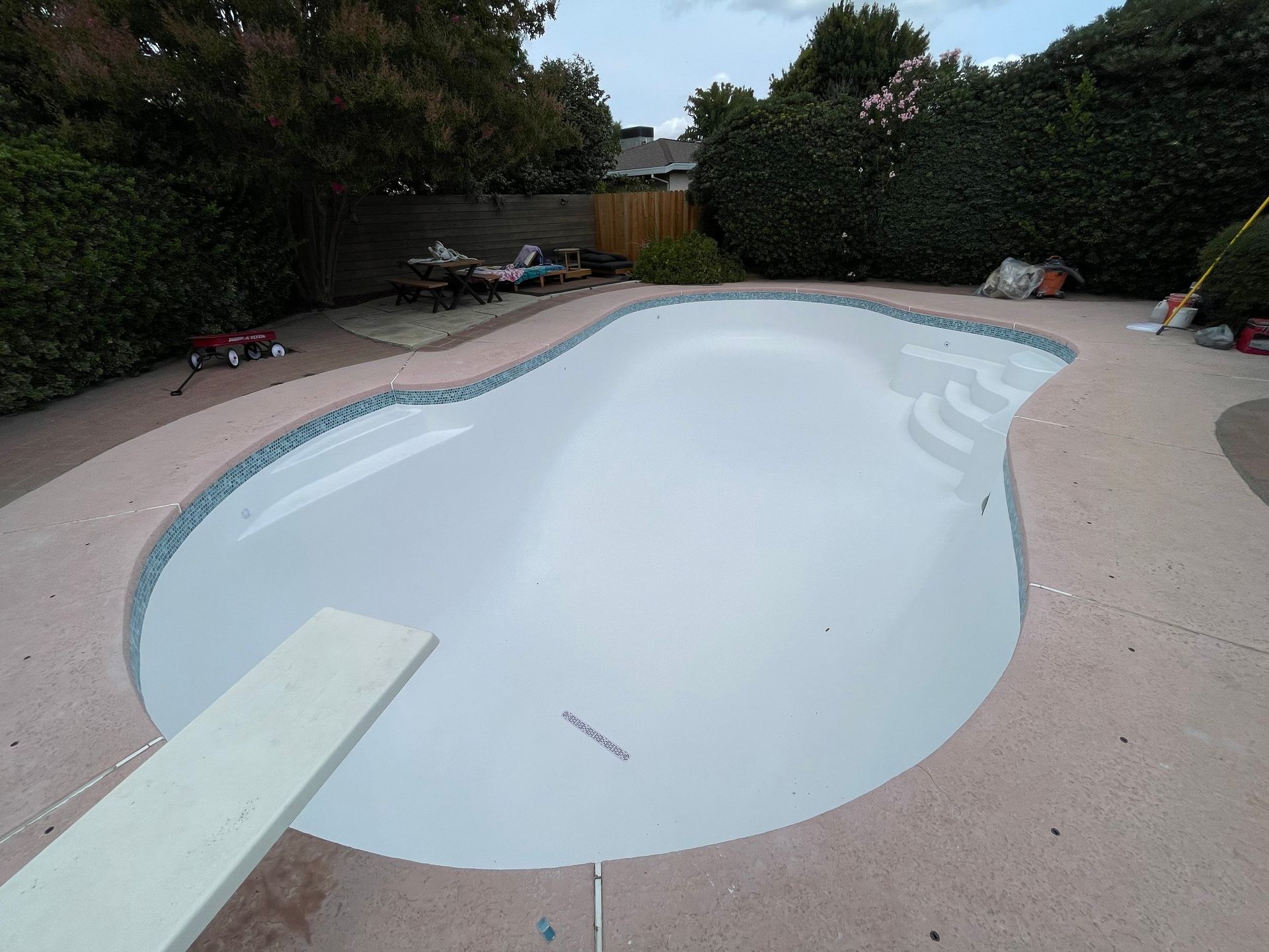 Empty kidney-shaped swimming pool with diving board, surrounded by concrete deck and landscaping. Empty kidney-shaped swimming pool with diving board, surrounded by concrete deck and landscaping.