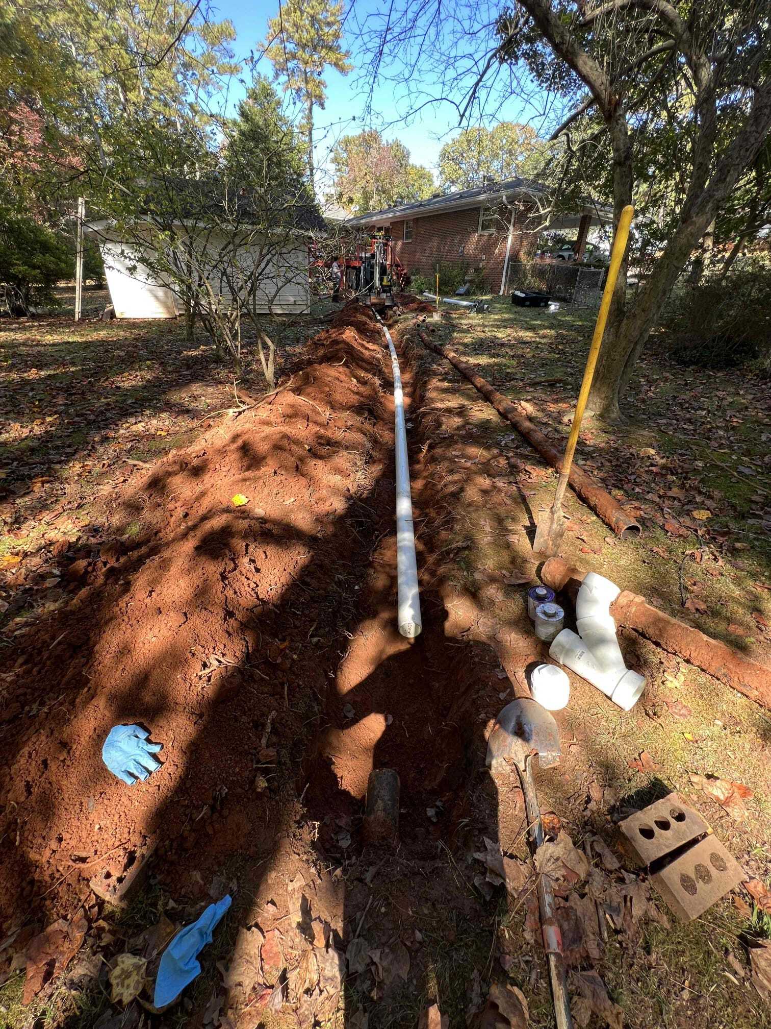 Trench dug in yard with white pipe laid inside, tools and supplies nearby, trees and house in background.
