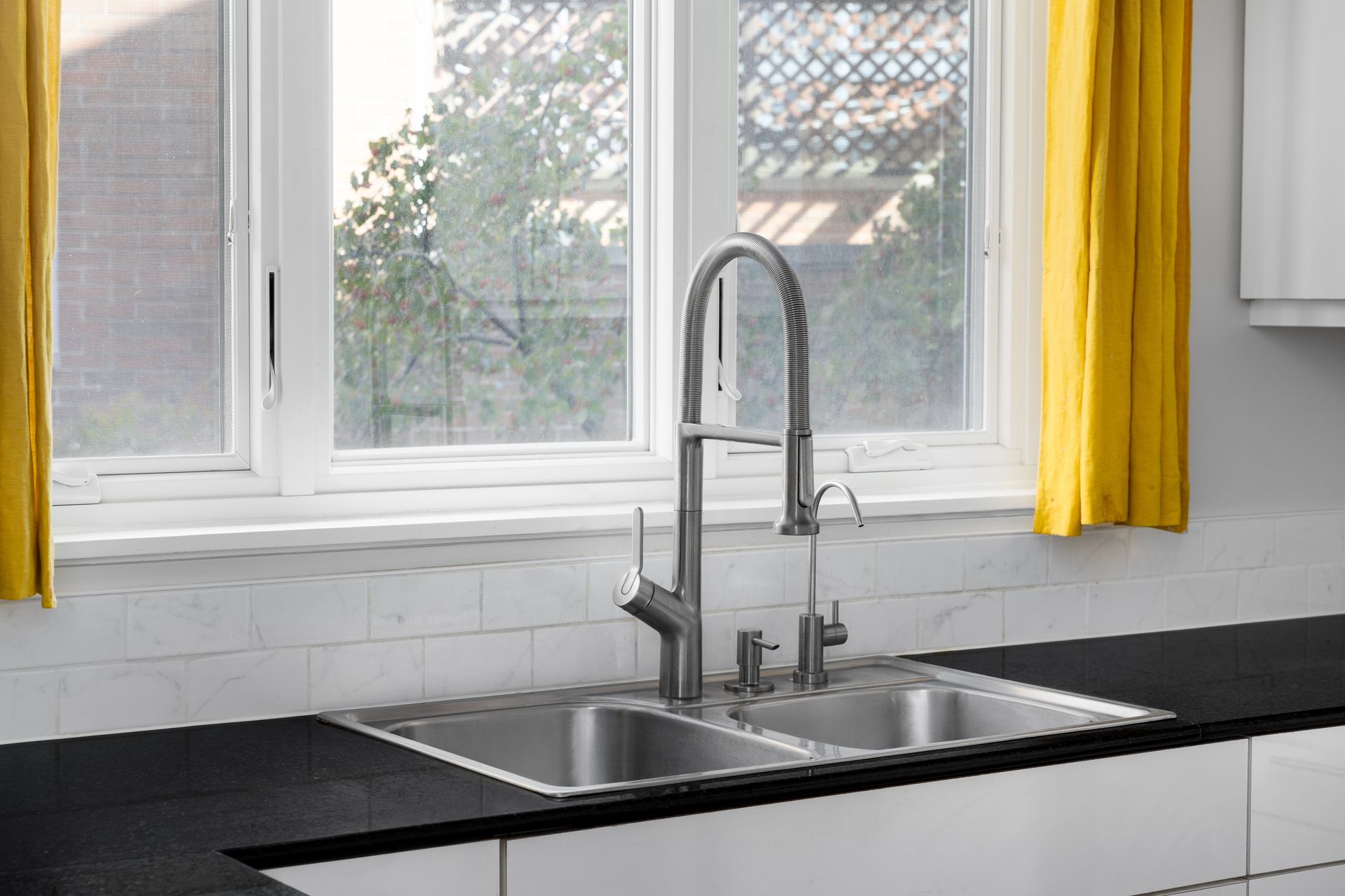 Kitchen sink with stainless steel double basin and a tall faucet. Bright yellow curtains frame the window above the sink.
