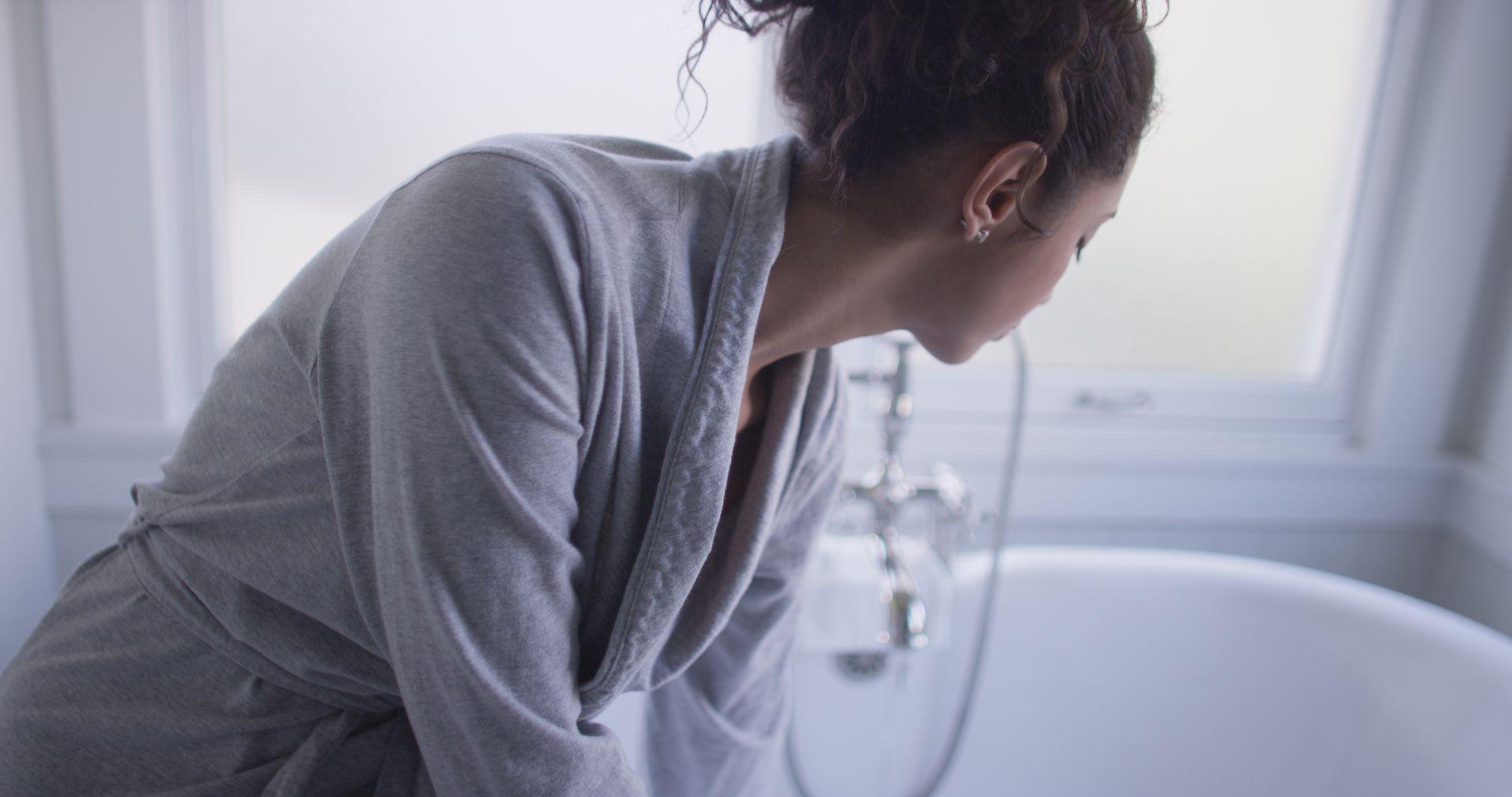 Woman in a grey robe leans over a white bathtub in a bathroom, looking down. A window is visible in the background.