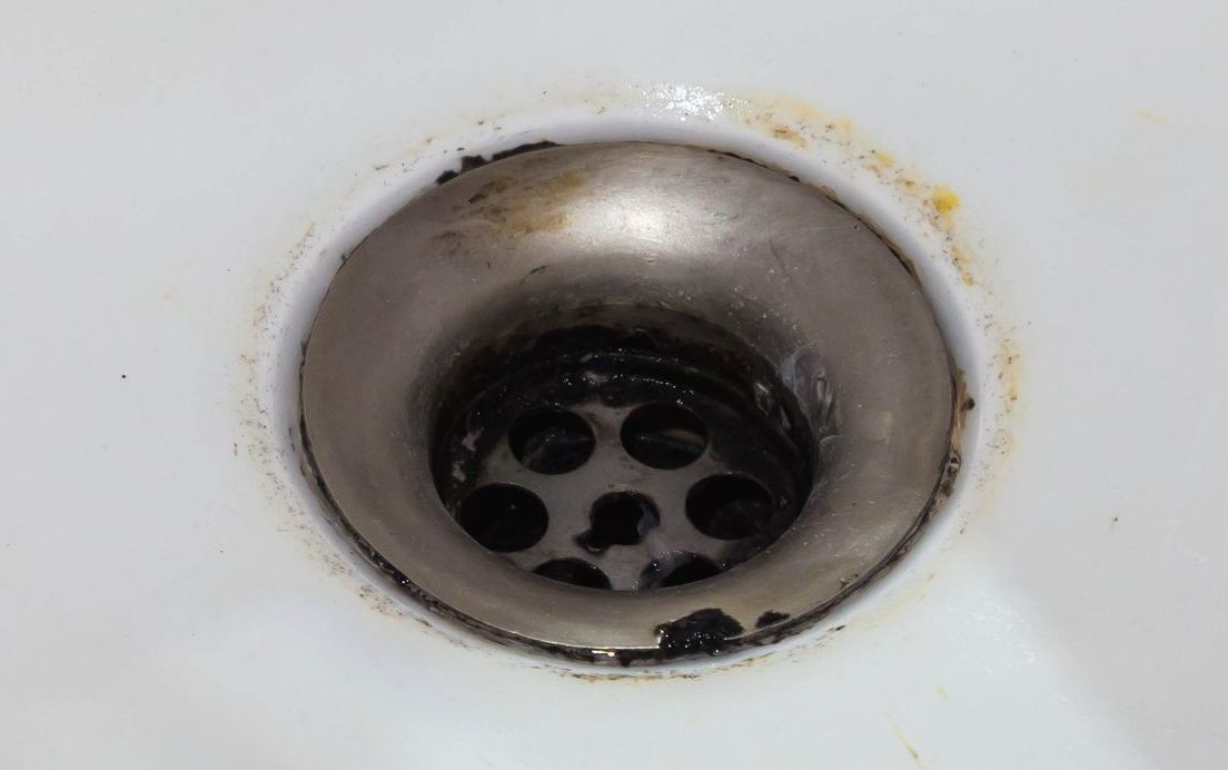 Close-up of a dirty sink drain with a metal strainer surrounded by white porcelain. The drain has black grime around its edges.