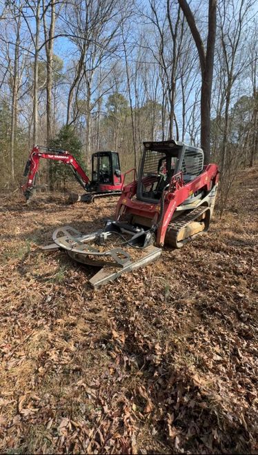 Red construction equipment, including a skid steer with a forestry cutter, parked in a wooded area covered in dry leaves.