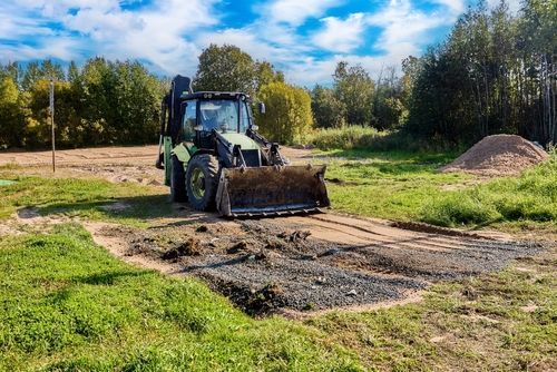 A green tractor with a front bucket parked on a dirt path in a grassy field near a forest and a pile of soil.