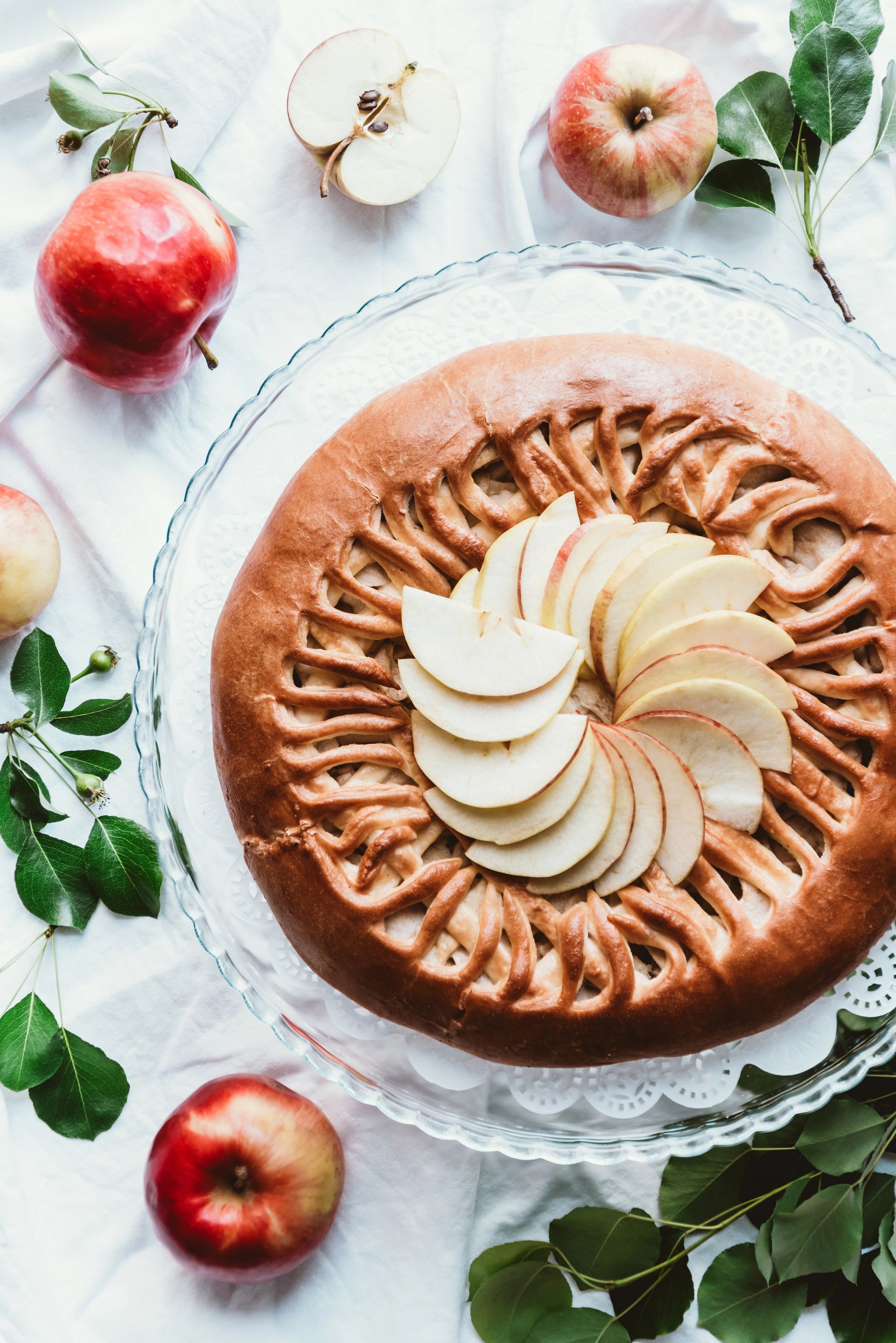 Torta di mele su un piatto di vetro, circondata da mele e verde; sfondo chiaro.