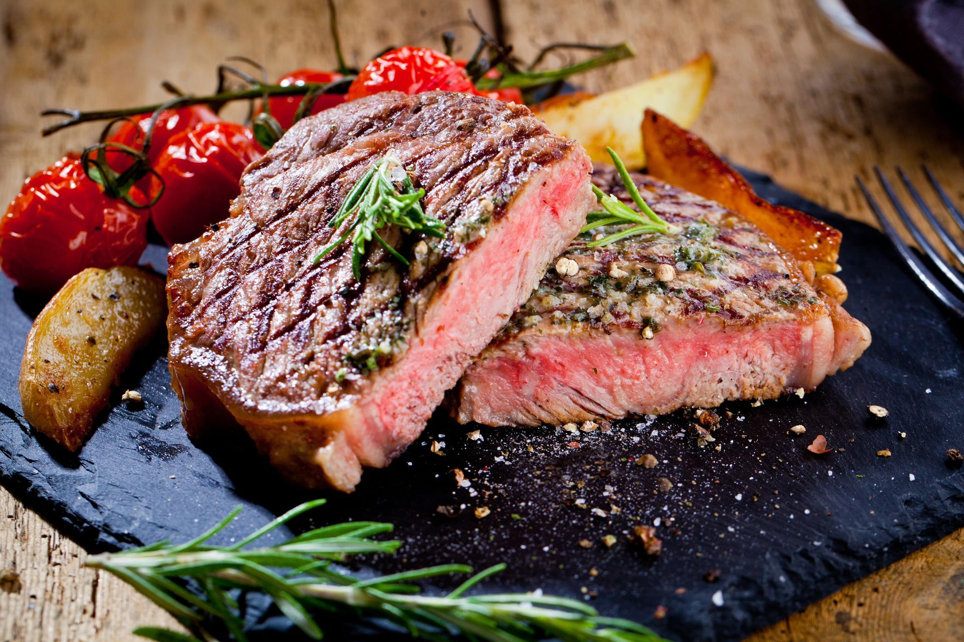 Bistecca tagliata a fette con erbe aromatiche, pomodori, patate e rametti di rosmarino su un tavolo di legno nero in ardesia.